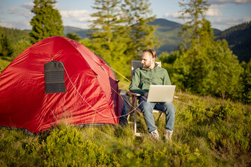 Man works on laptop while traveling with tent in the mountains. Charging computer with portable...