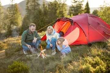 Young adult couple sit with a little girl by the campfire at campsite, traveling with tent on...
