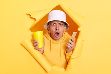 Horizontal shot of amazed surprised man wearing hoodie and panama breaking through torn in yellow...