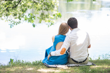 A couple in love sits on the shore of a lake under a tree. Hugs of a man and a woman at a picnic. Love story or valentine's day concept. View from the back of couple sitting in summer park or forest