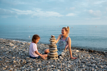 A mother and daughter 6 years old on the beach build a castle of stones. They socialize and have fun together. Family. Summer. Vacation.