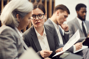 Attractive young businesswoman in glasses sitting in auditorium and talking to colleague about application form