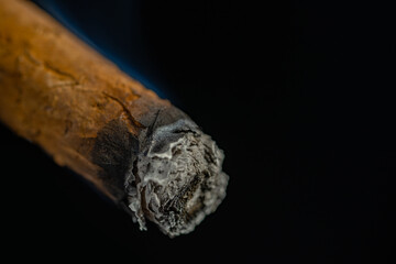 Smoking cuban cigar with blue smoke on an isolated black background. A Havana cigar burns and emits smoke in a dark bar. A rolled cigarette made from tobacco leaves. Macro shot of a burning cigar.