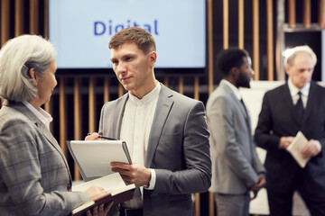 Serious handsome young journalist in suit making notes in clipboard while interviewing business forum participant during break