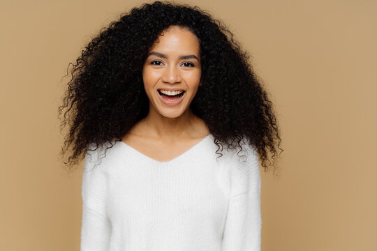 Headshot of healthy positive female model laughs happily, has bushy Afro hair, dressed in white soft jumper, glad to receive compliment from boyfriend, isolated over brown background. Emotions concept