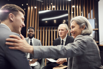 Smiling mature Asian businesswoman with gray hair sitting among colleagues in conference room and touching shoulder of young specialist while celebrating him with success