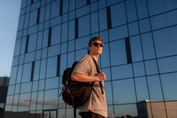 Portrait of handsome tourist hipster man in sunglasses with bag handy, standing guy near the building in downtown. Calm relaxing moment, summer vacation concept - 516445154