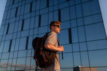 Portrait of handsome tourist hipster man in sunglasses with bag handy, standing guy near the building in downtown. Calm relaxing moment, summer vacation concept - 516445302