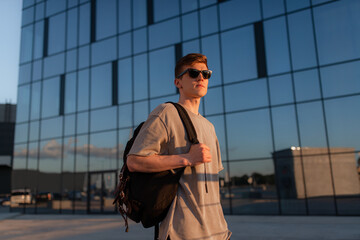 Portrait of handsome tourist hipster man in sunglasses with bag handy, standing guy near the building in downtown. Calm relaxing moment, summer vacation concept - 516445558