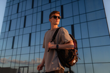 Portrait of handsome tourist hipster man in sunglasses with bag handy, standing guy near the building in downtown. Calm relaxing moment, summer vacation concept - 516445941