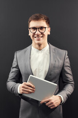 Portrait of happy intelligent young entrepreneur in glasses standing against black background and holding digital tablet