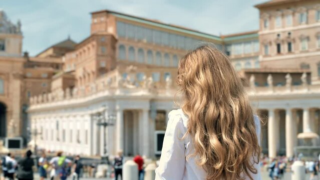View of the blonde woman from behind in Vatican City. Saint Peter Square with multiple tourists, Saint Peter Basilica and the Vatican Obelisk on the background. Slow motion