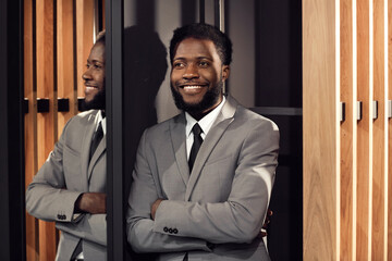Smiling satisfied young African-American manager with beard leaning on wall and observing business meeting