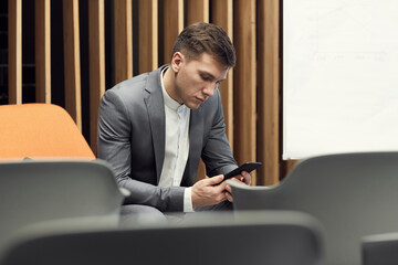 Serious handsome young businessman in gray suit sitting in empty conference room and using smartphone