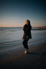 A woman enjoys the sunset at the seaside on a cold day. Portrait of a young lady on the beach during sunset. Lady walks on a cold evening