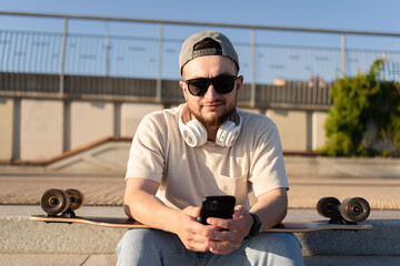 Young man using his smartphone for listening to music. Skater relax at the street. Hipster man skateboarding walking in the city alone.