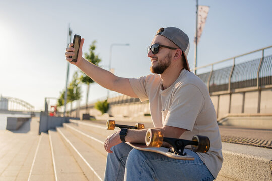 Young hipster man taking photo with his phone sitting with the longboard.