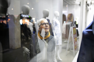 Smiling mature senior woman with short gray hair and eyeglasses looking in the shop window, night scene