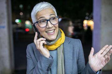 Smiling mature senior woman with short gray hair and eyeglasses use phone on street, night scene in city