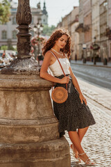 Elegant curly brunette woman wearing trendy summer outfit with round wicker shoulder rotan bag,...
