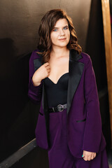 a brunette woman in a lilac suit and a sexy black top against a black wall 