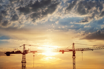 Scenic view of two high tower crane silhouette working at early sunrise time at construction site against bright orange dawn sun light cloud sky. City urban development concept - Powered by Adobe