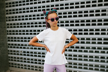 Naklejka premium Portrait of a cute teenage girl with African braids in a white t-shirt and sunglasses stands on a city street. Mock-up for print. T-shirt template.