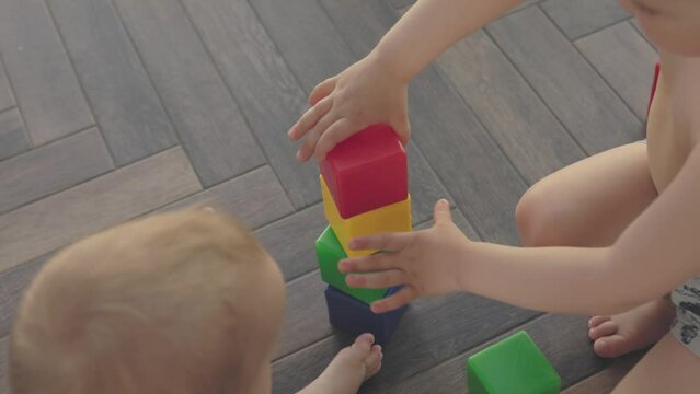 two kids building tower from colorful plastic cubes together on brown floor in room. destroying crashing building during children game. siblings play together, toddler child helping brother kid put 