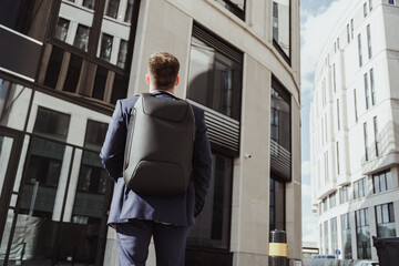 Young clerk in suit with backpack stands in front of corporate building and looks up, back view