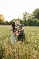 Cheerful young parents embracing little cute daughter standing outdoor. Caucasian family of three, smiling father kissing mother and holding little baby girl on summer nature.