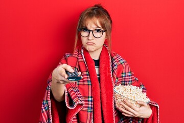 Redhead young woman eating popcorn using tv control depressed and worry for distress, crying angry...