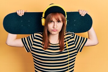 Redhead young woman holding skate wearing headphones skeptic and nervous, frowning upset because of...