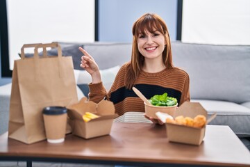 Young beautiful woman eating delivery food at the living room smiling happy pointing with hand and finger to the side