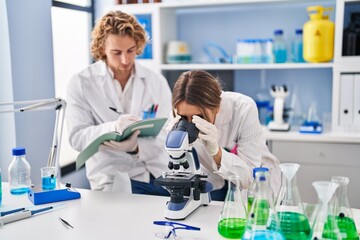 Fototapeta premium Man and woman wearing scientist uniform writing on notebook using microscope at laboratory