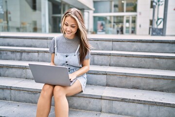 Young caucasian girl smiling happy using laptop sitting on the stairs at the city.