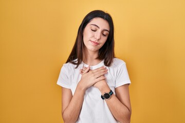 Young beautiful woman standing over yellow background smiling with hands on chest with closed eyes and grateful gesture on face. health concept.