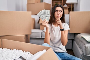 Young beautiful woman sitting on the floor at new home holding money serious face thinking about...