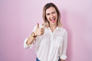 Young beautiful woman standing over pink background doing happy thumbs up gesture with hand. approving expression looking at the camera showing success.