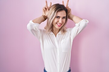 Young beautiful woman standing over pink background posing funny and crazy with fingers on head as bunny ears, smiling cheerful