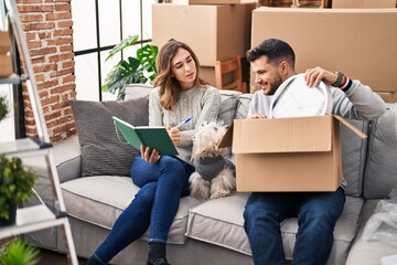 Man and woman unpacking box writing on notebook sitting on sofa with dog at new home