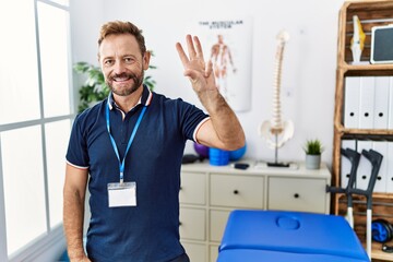 Middle age physiotherapist man working at pain recovery clinic showing and pointing up with fingers number three while smiling confident and happy.