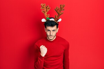 Young hispanic man wearing cute christmas reindeer horns angry and mad raising fist frustrated and...