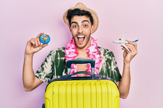 Young hispanic man wearing summer style and hawaiian lei holding world ball and plane toy celebrating crazy and amazed for success with open eyes screaming excited.