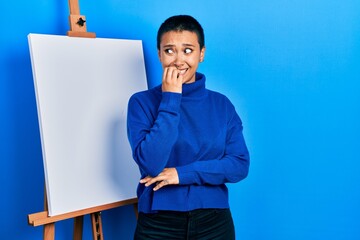 Beautiful hispanic woman with short hair standing by painter easel stand looking stressed and...
