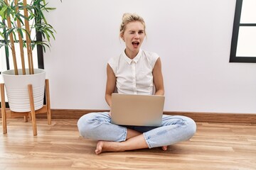 Young blonde woman using computer laptop sitting on the floor at the living room winking looking at...