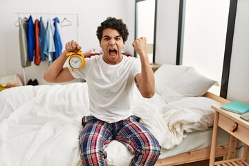 Young hispanic man holding alarm clock sitting on the bedroom annoyed and frustrated shouting with...