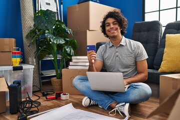 Young hispanic man using laptop and credit card sitting on floor at new home