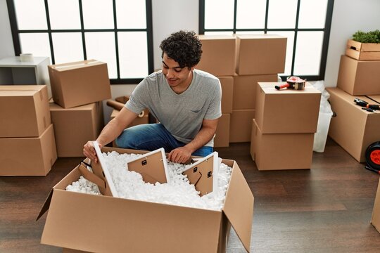 Young hispanic man smiling happy unboxing cardboard box at new home.
