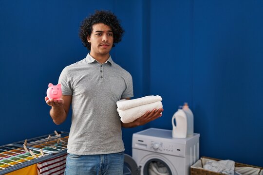 Hispanic man with curly hair holding clean towels and piggy bank skeptic and nervous, frowning upset because of problem. negative person.