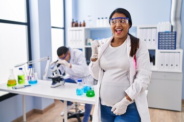 Fototapeta premium Young hispanic woman expecting a baby working at scientist laboratory smiling with happy face looking and pointing to the side with thumb up.